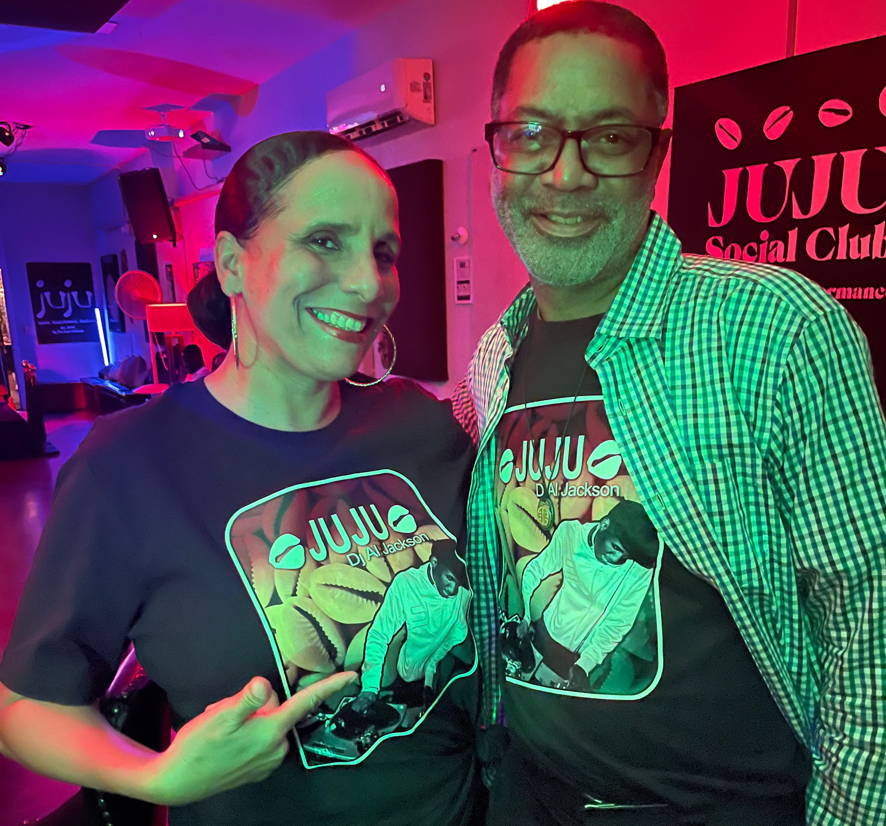 Two people wearing 'Juju' branded shirts in a social club setting with colorful lighting.